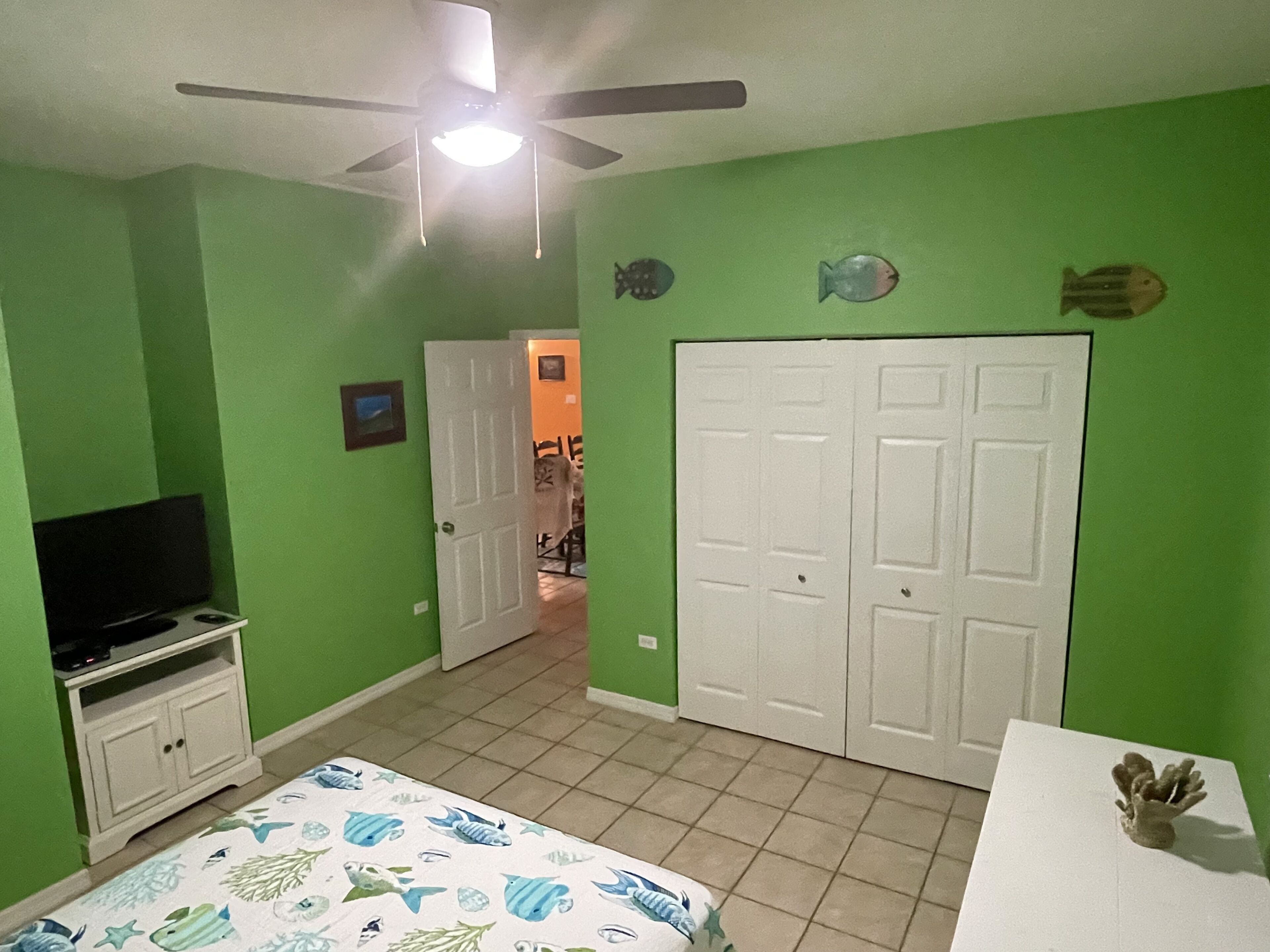 Een kamer met groene wanden, een plafondventilator, een bed met een sprei met een vispatroon, een televisie op een standaard en een witte deur.