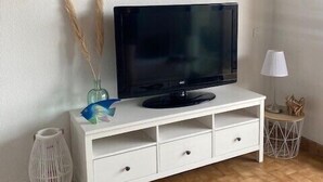 Un meuble blanc pour la télévision, sur lequel est posée une télévision à écran plat, un tableau décoratif au-dessus, et une petite table avec une lampe sur le côté droit.