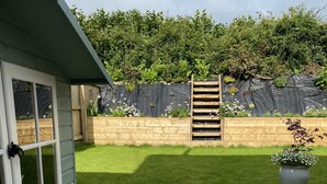 Un espace patio avec une terrasse en bois, un ensemble de marches en béton menant à une porte, et un jardin avec une clôture en bois et des plantes.