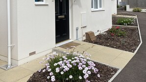 Une maison à deux étages avec une porte d’entrée noire, des moulures blanches et un petit porche. Devant la maison, on trouve un parterre de fleurs avec des fleurs roses.