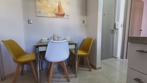 A dining area with a table set for two, two yellow chairs, a white chair, and a painting of a sailboat on the wall. - Mon Repo Nest (Corfu)