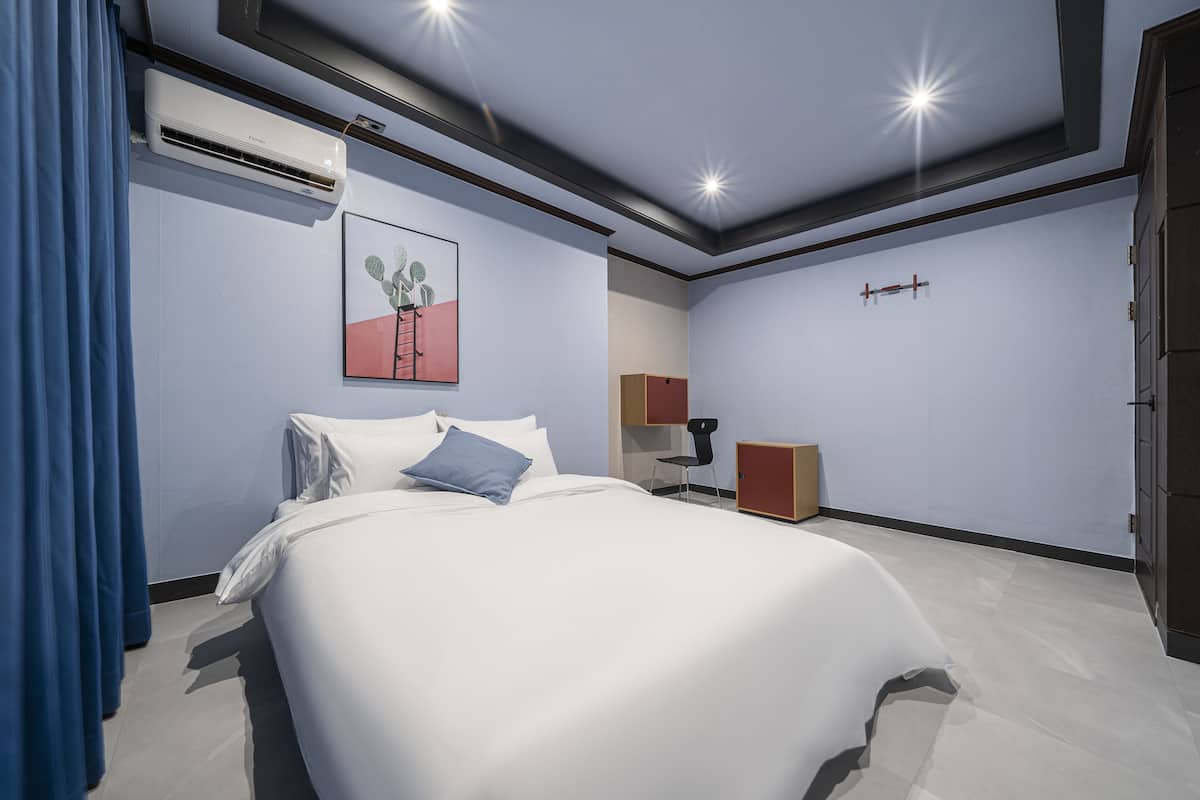 A hotel room with a bed, a desk, a chair, an air conditioning unit, and a door.