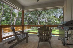 Ein überdachter Außenbereich mit zwei Adirondack-Stühlen, einem Grill und Blick auf ein bewaldetes Gebiet.