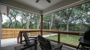 A covered outdoor area with a wooden deck, two Adirondack chairs, a grill, and a view of trees and a fence.