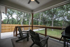 Un espacio al aire libre techado con una terraza de madera, dos sillas Adirondack, una parrilla y vistas a árboles y una valla.