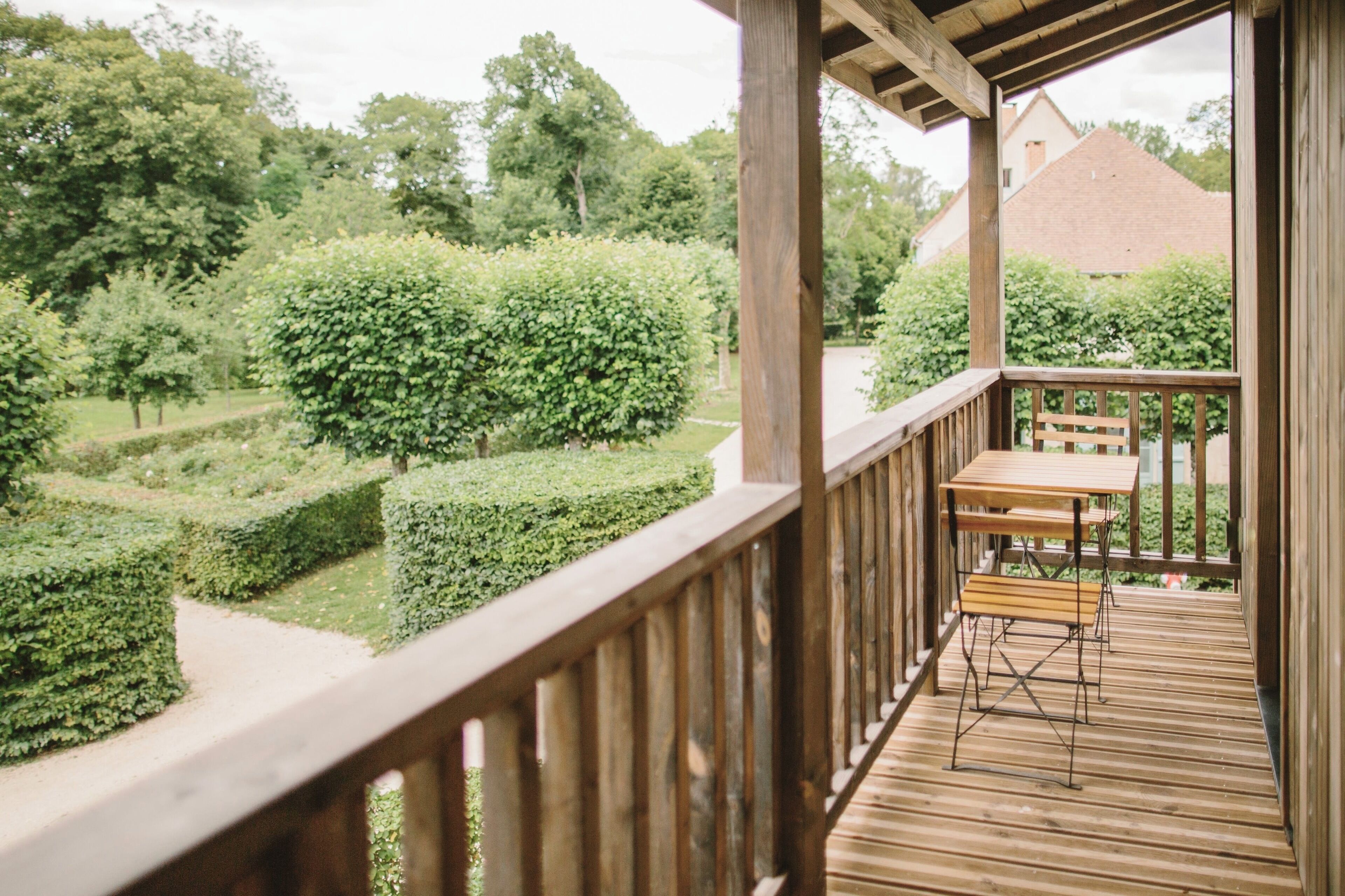A wooden deck with a table and bench, overlooking a well-manicured garden with hedges and trees.