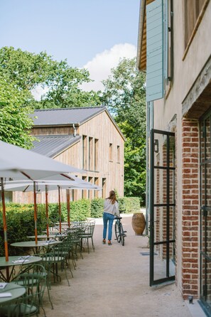 An outdoor seating area with tables and chairs, a person with a bicycle, and a building with a brick facade. - Domaine de Primard - Fontenille Collection (Guainville)