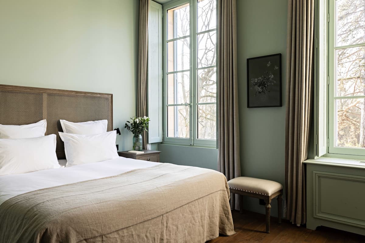 A bedroom with a large bed, a nightstand, a bench, and a window with curtains.