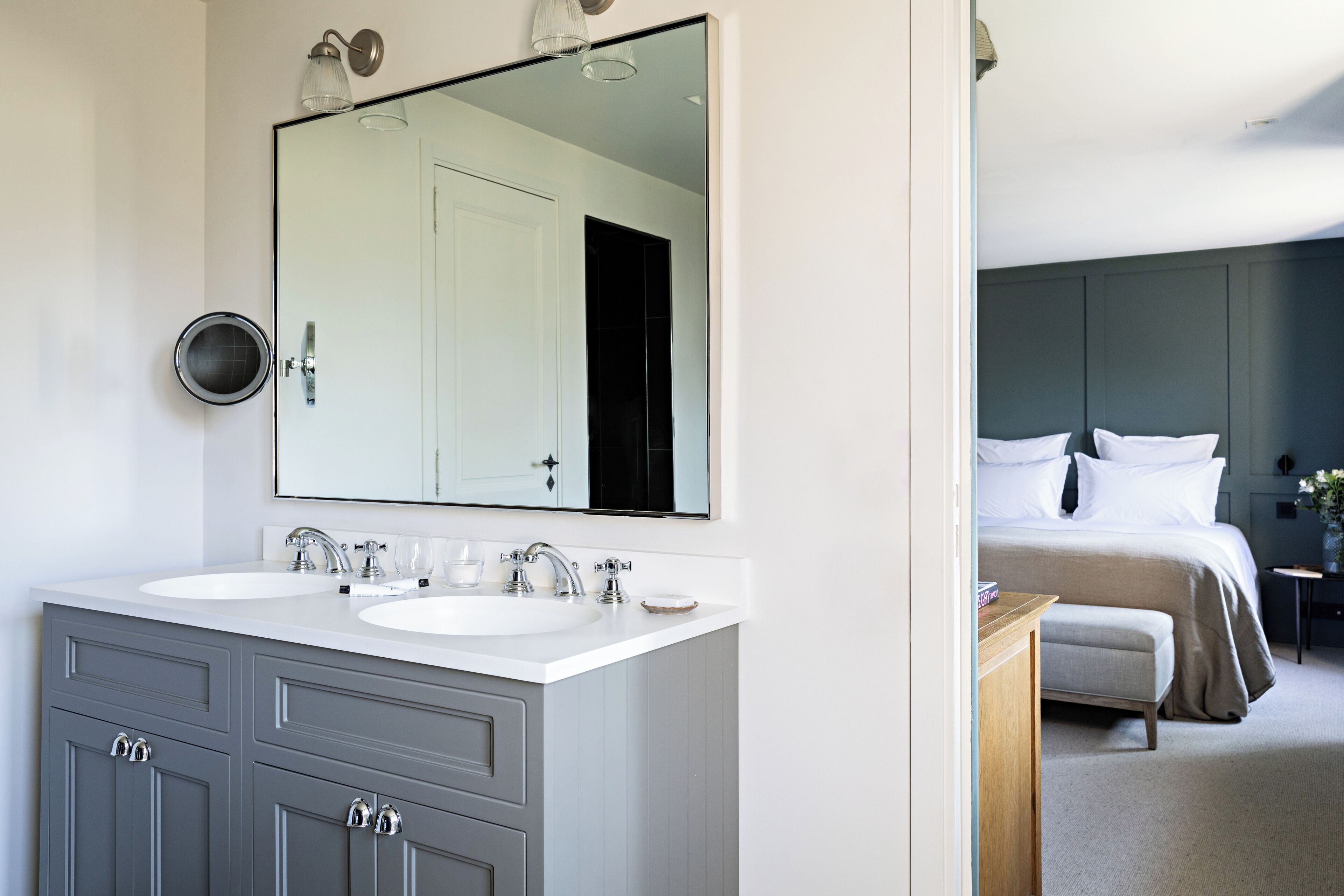 A modern bathroom with dual sinks, a large mirror, and a dark-toned bedroom with a bed and bedside table.