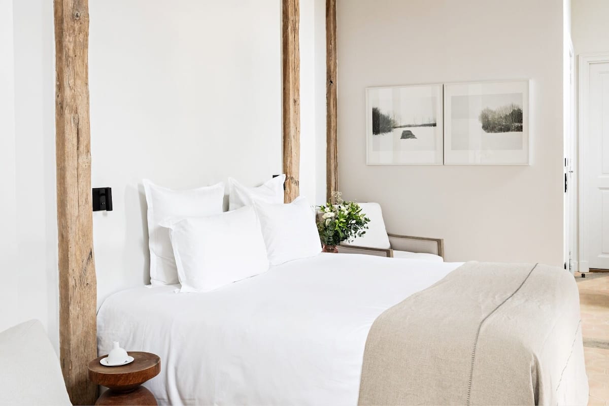 A minimalist bedroom with a large bed, white bedding, and a small bedside table with a lamp.