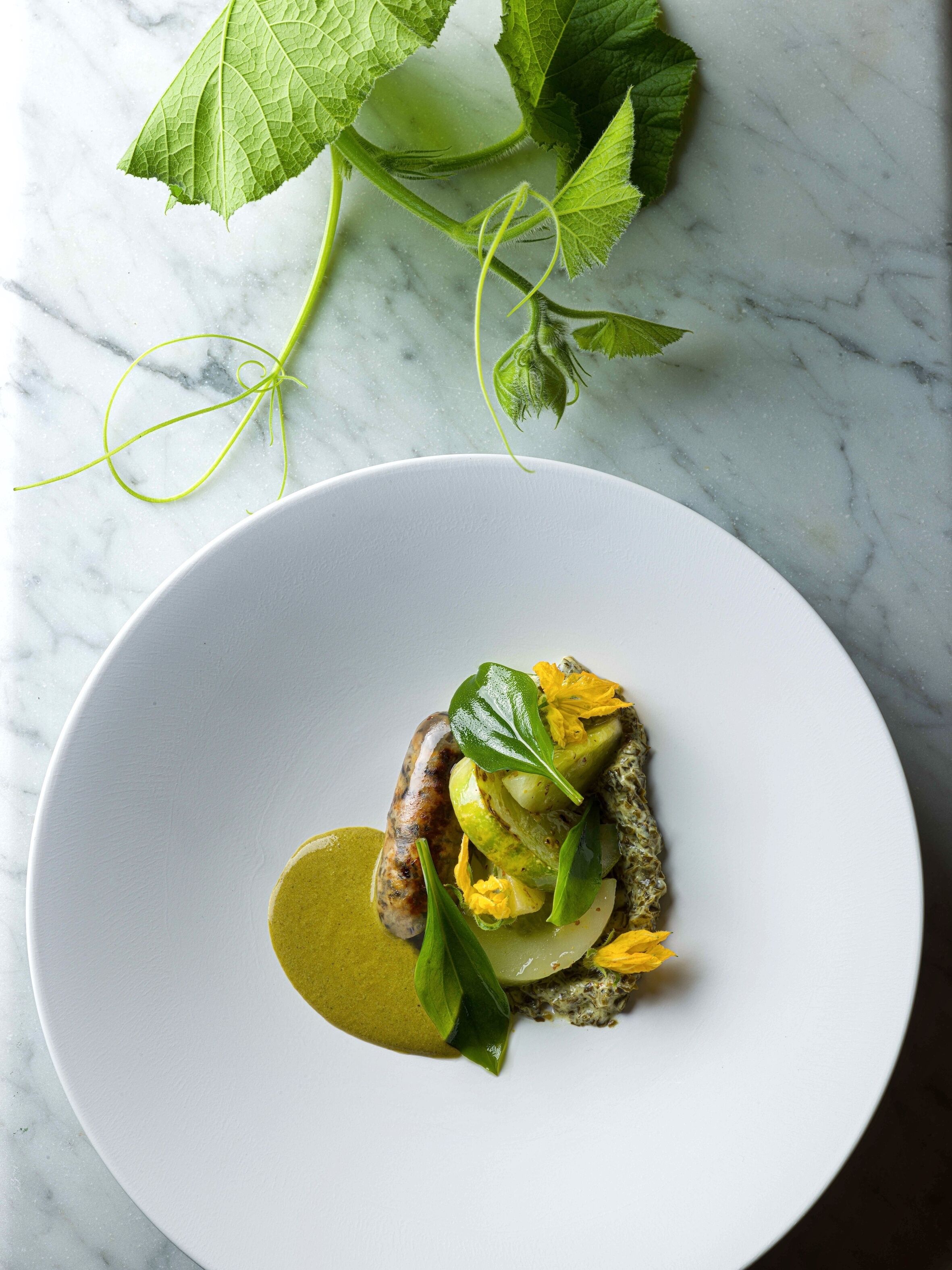 Une assiette blanche contenant un plat composé d’une saucisse, de légumes et d’une sauce verte, le tout posé sur une surface en marbre.