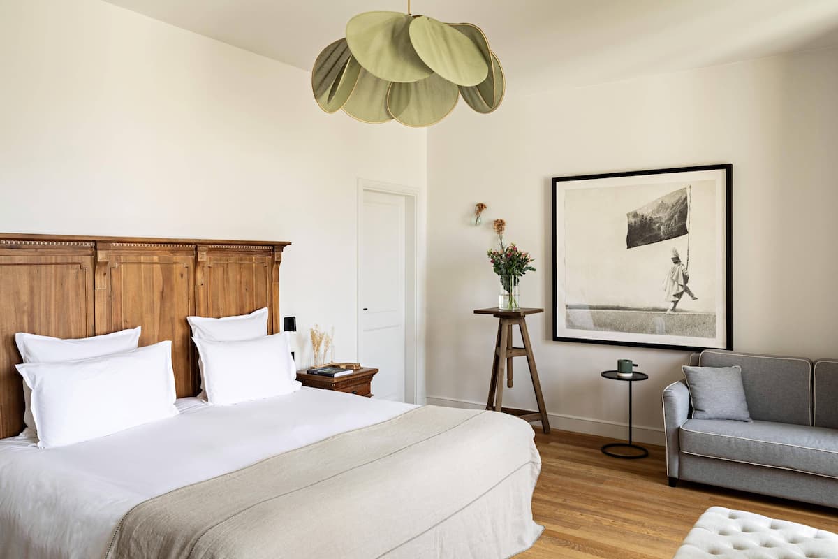 A bedroom with a large bed, a wooden headboard, a grey sofa, a small side table, a framed picture on the wall, and a hanging light fixture.