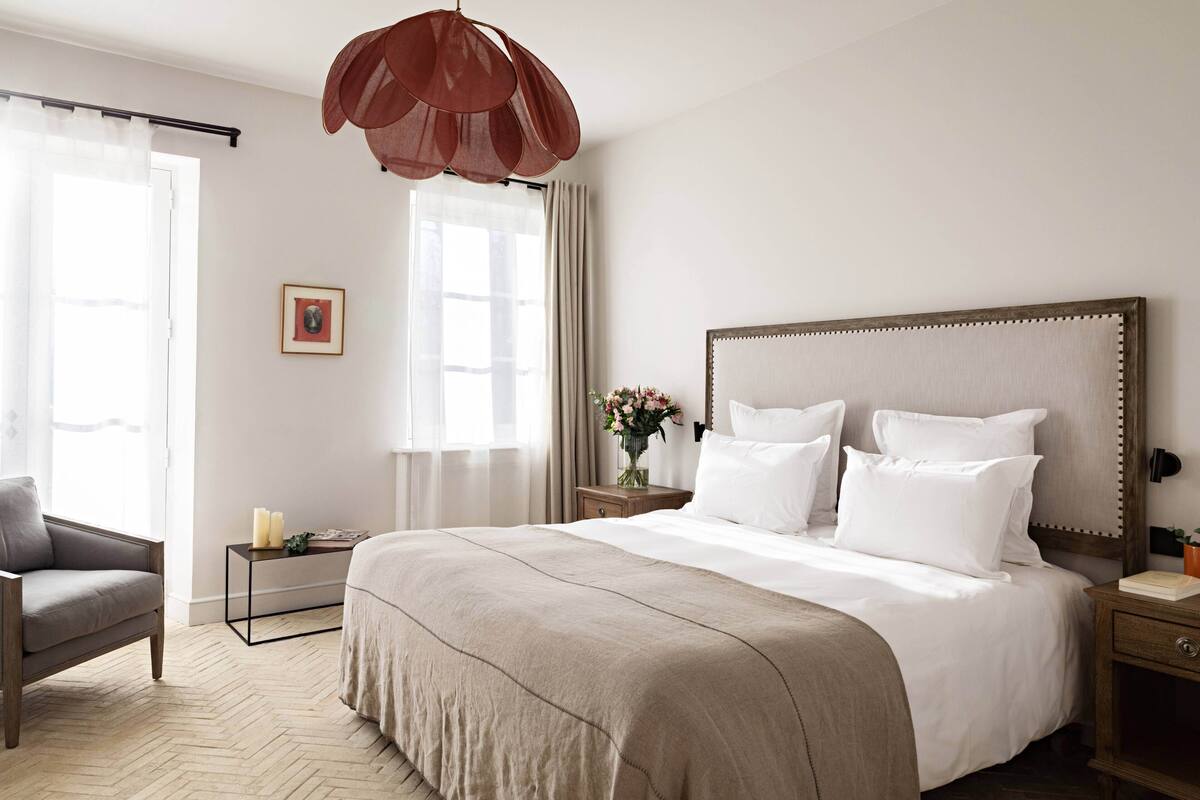 A bedroom with a large bed, a bedside table with a vase of flowers, a chair, and a large window with curtains.