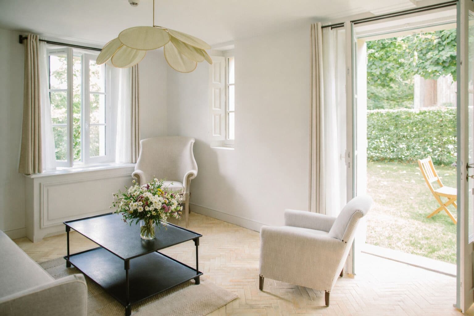 A spacious room with a large window, a white armchair, a black coffee table with a floral arrangement, and a hanging light fixture.