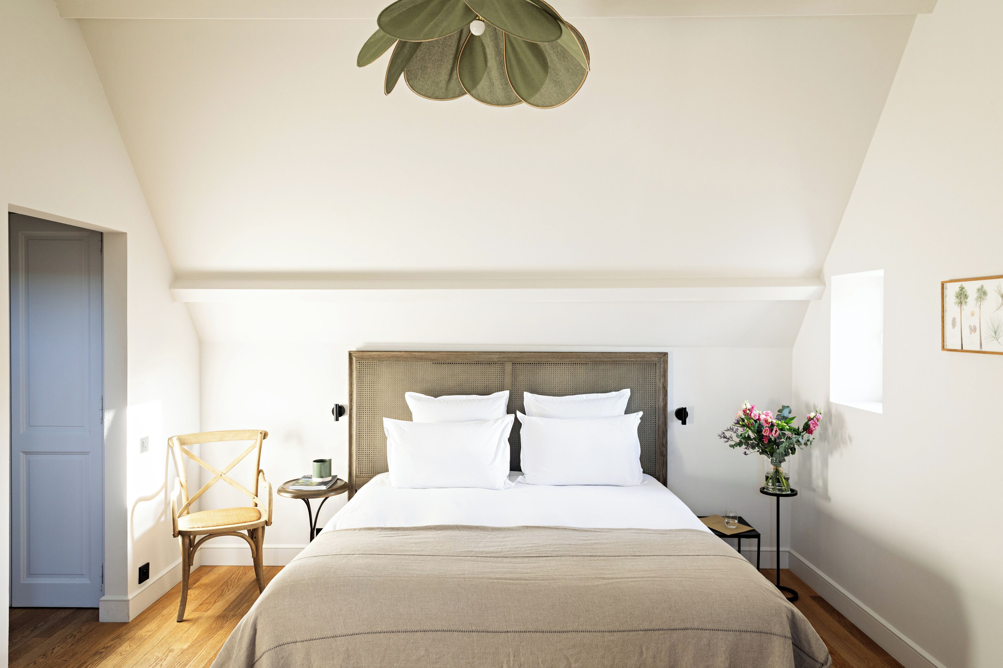Un dormitorio con cabecera de madera, cama con sábanas blancas, una silla de madera, una mesita con un jarrón de flores y una puerta a la izquierda.