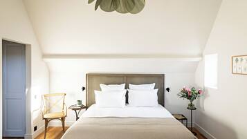 Un dormitorio con cabecera de madera, cama con sábanas blancas, una silla de madera, una mesita con un jarrón de flores y una puerta a la izquierda.