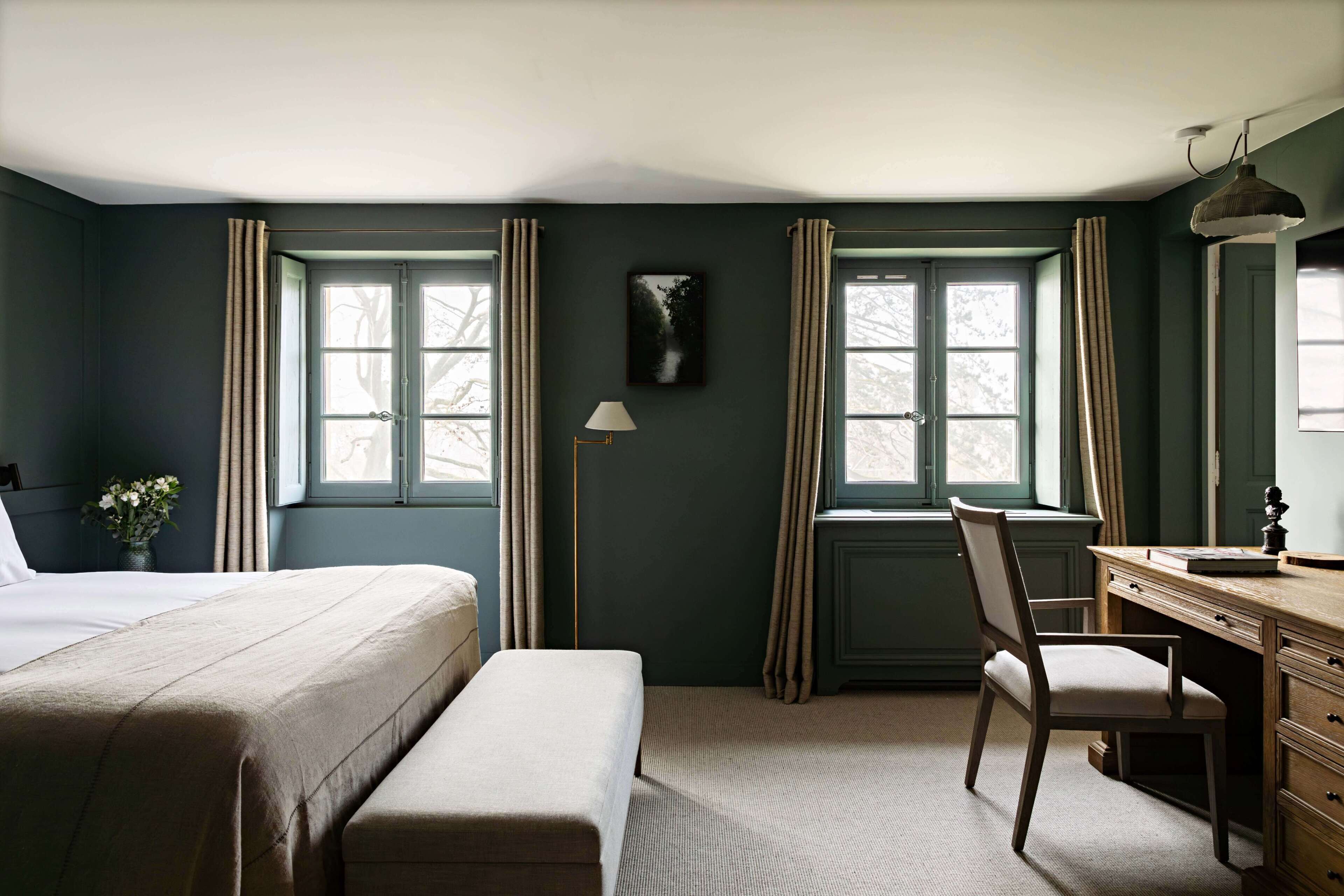 Une pièce comprenant un lit, un bureau et une chaise. Il y a des fenêtres avec des rideaux, une lampe et un tableau au mur.