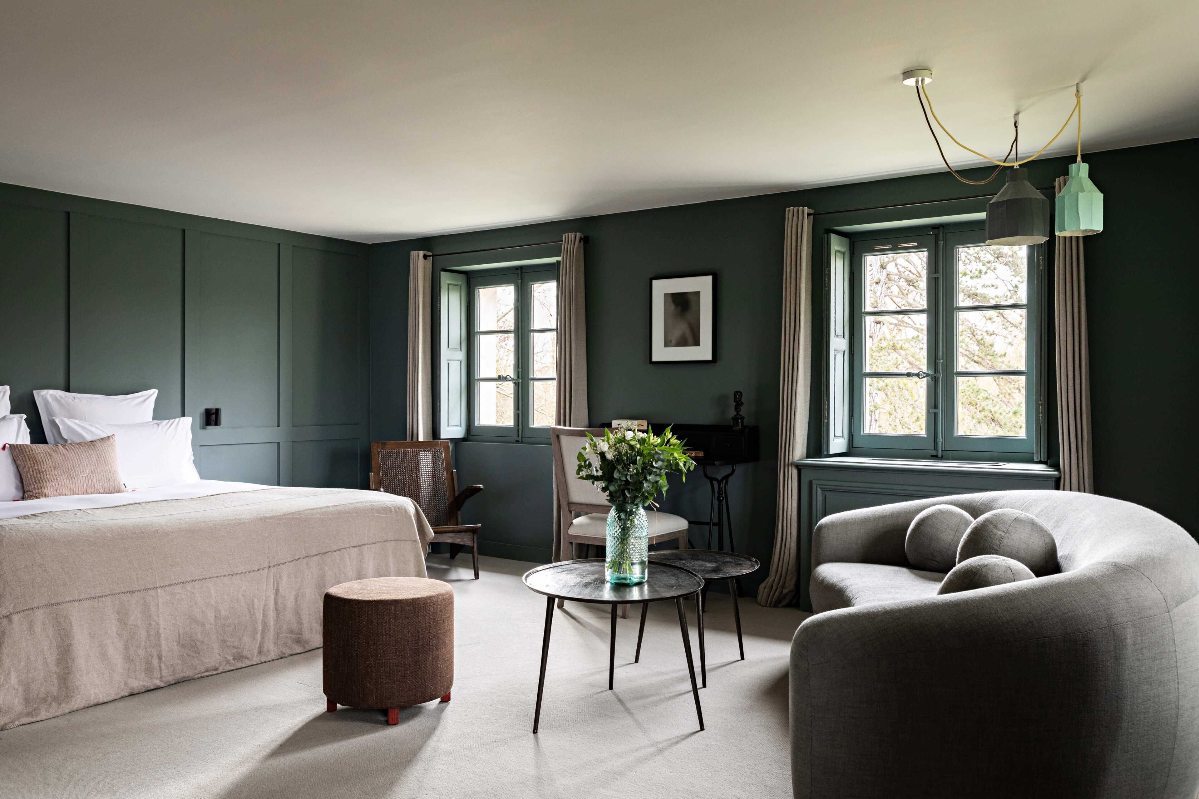 A room with a bed, two chairs, a small table with a vase, and a sofa.