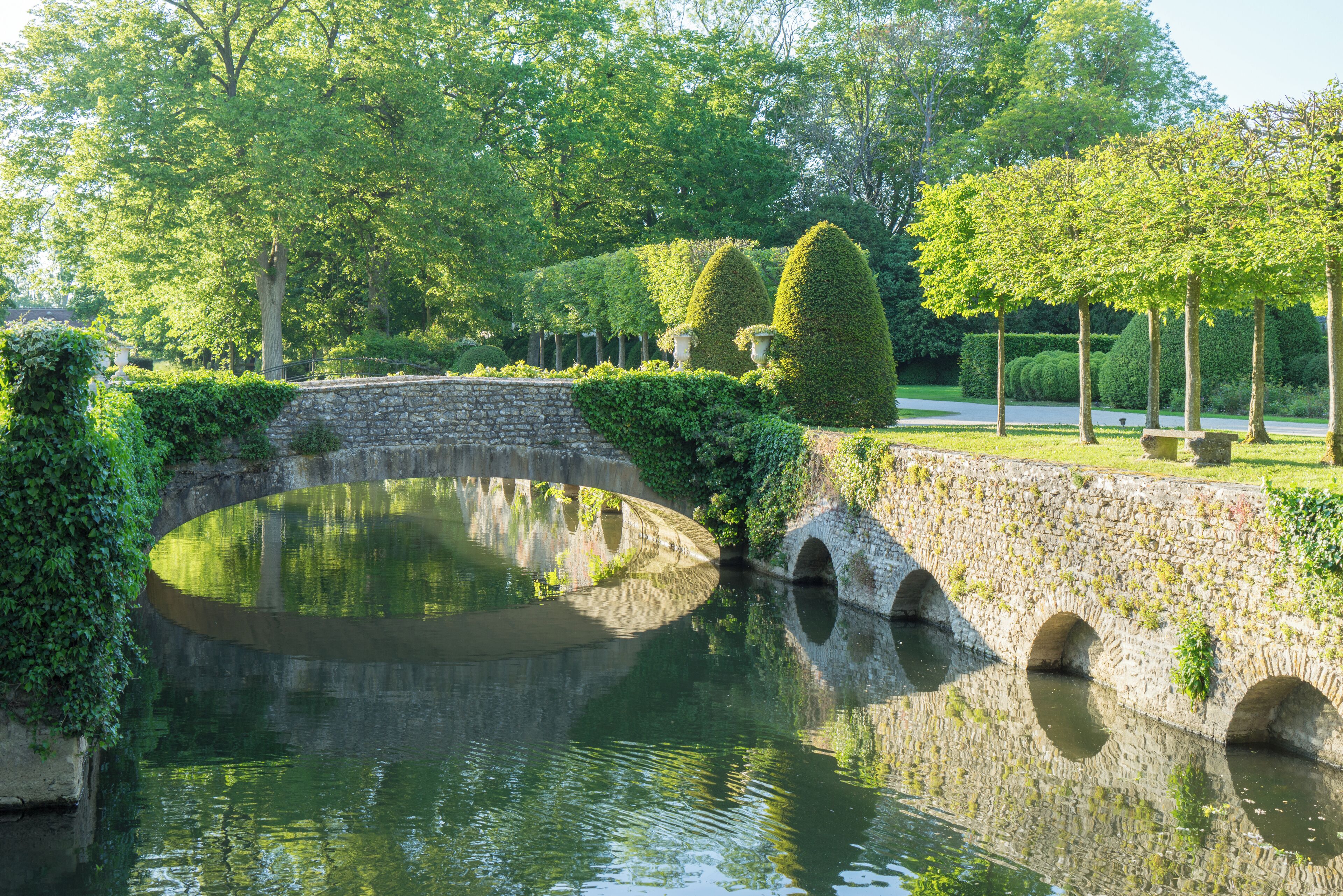 A stone bridge over a calm water body, surrounded by lush greenery and trees.