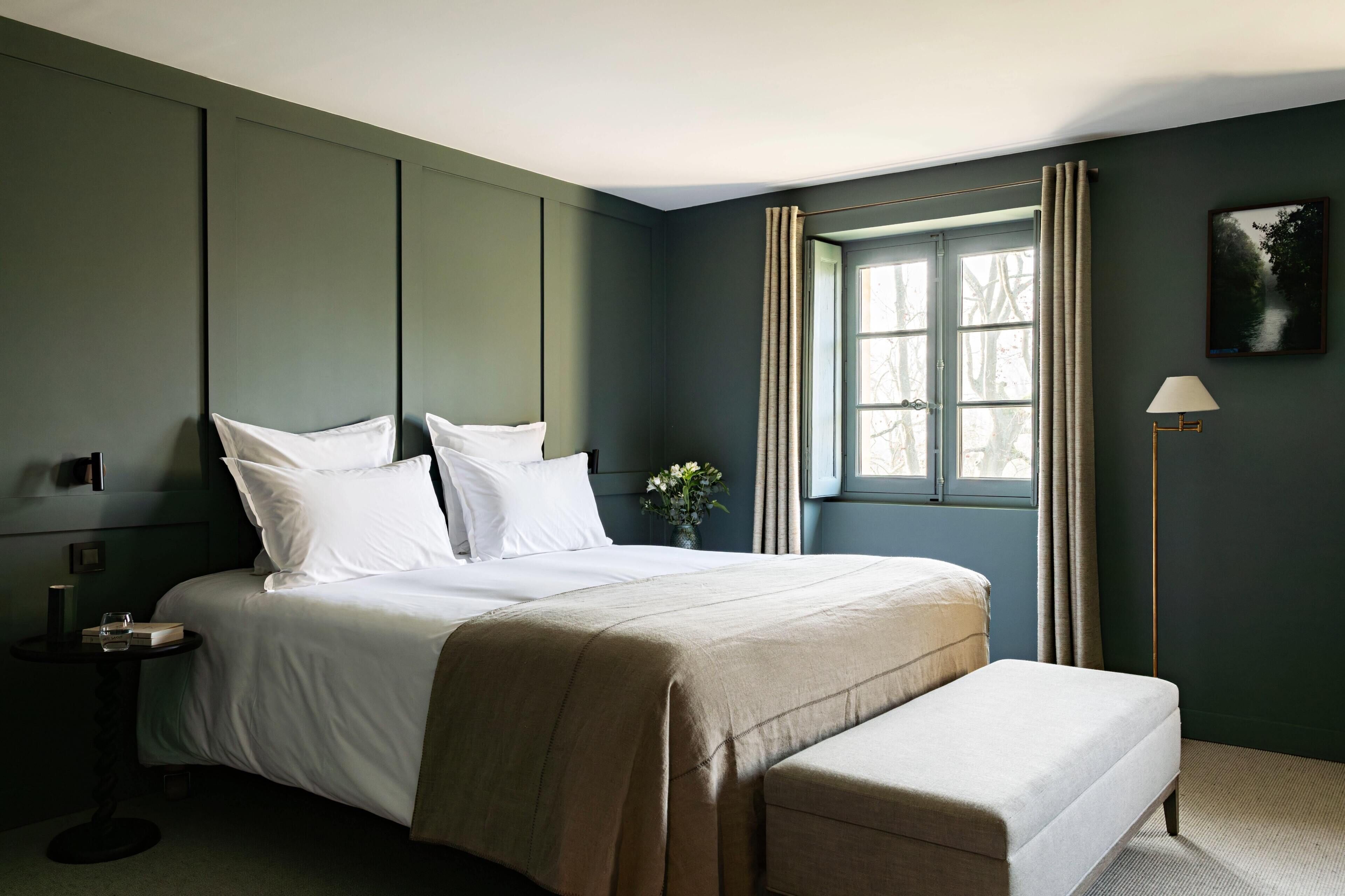 Una cama bien hecha con sábanas blancas y una manta color beige, una mesita de noche con lámpara, y una ventana con cortinas.