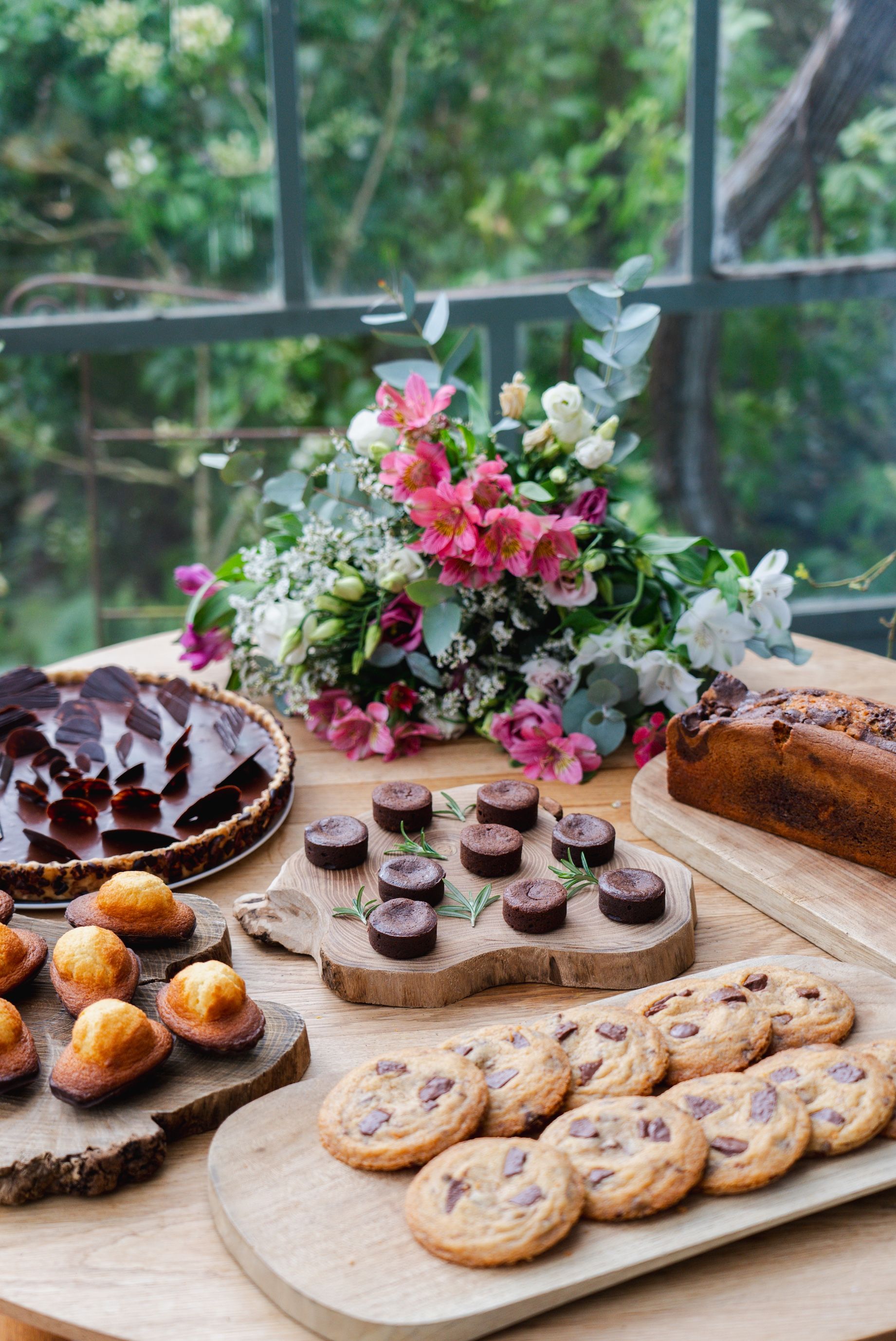 Verschiedene gebackene Köstlichkeiten auf einer Holzplatte, darunter Kekse und Muffins, mit einer Blumenkomposition im Hintergrund.