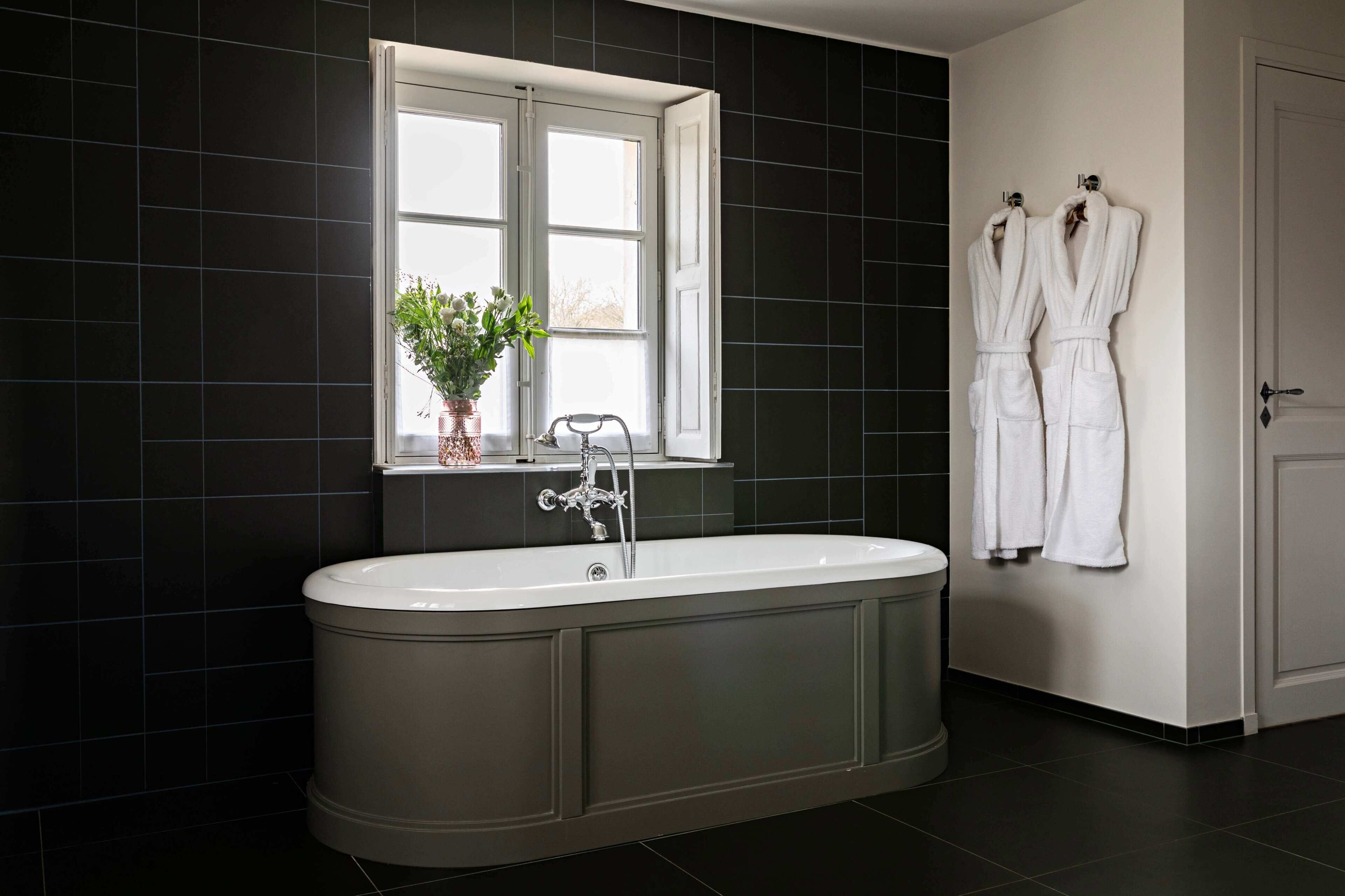 Een moderne badkamer met een vrijstaand bad, een raam met een plant in pot, en twee witte badjassen hangend aan de muur.