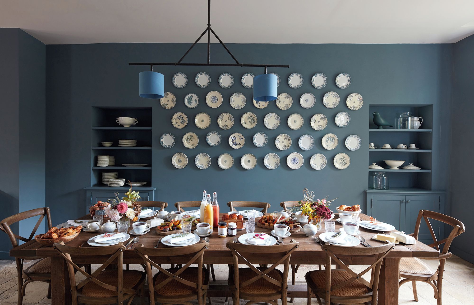 A dining room with a table set for breakfast, surrounded by chairs, with a wall of plates above.