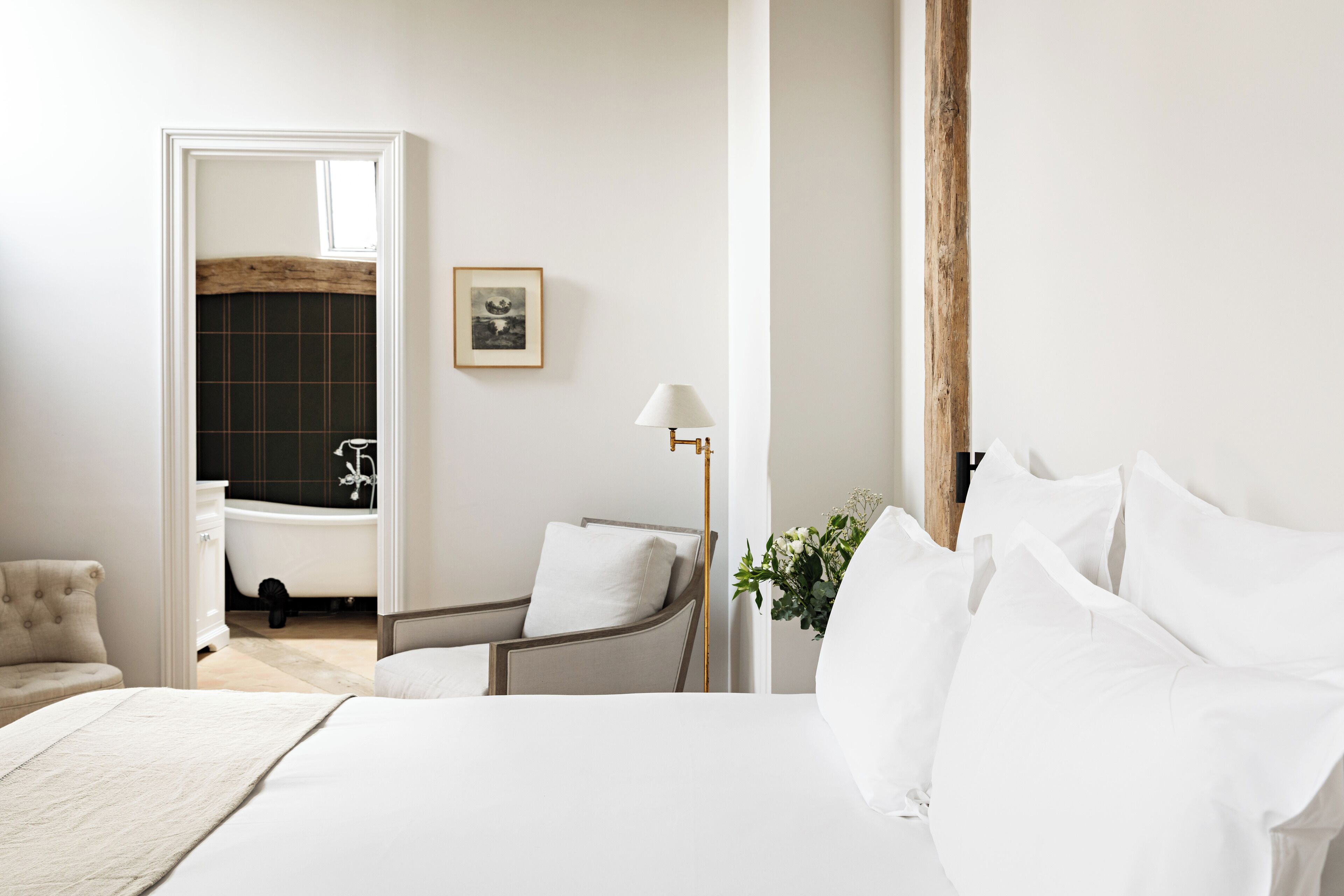 A bedroom with a bed, a chair, a bedside table, a lamp, and a bathroom visible through an open door.