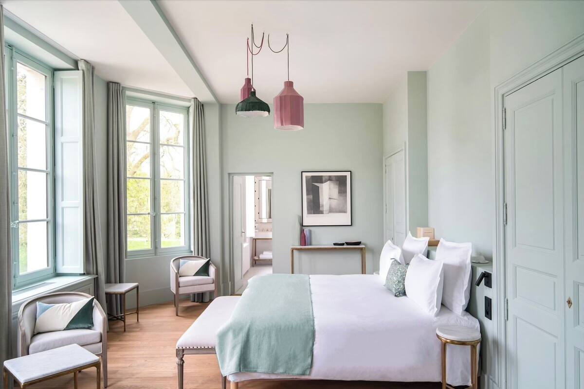 A bedroom with a large bed, bedside tables, a chair, and a desk. There are windows with curtains, a ceiling light, and a framed picture on the wall.
