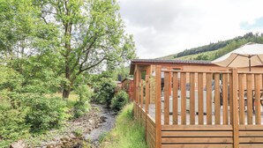 Une cabane en bois dotée d’une terrasse donnant sur un ruisseau, entourée d’une végétation luxuriante et d’arbres.