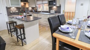A modern kitchen with a central island, bar stools, a dining table set for two, and a view of the outside. - Lavender Lodge (Windermere)