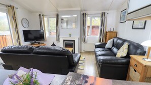 A modern living room with a black leather sofa, a flat-screen TV, a fireplace, and a view of the outdoors through large windows. - Lavender Lodge (Windermere)