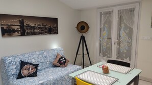 Une pièce avec un canapé à motifs, une table à manger ornée d’un chemin de table à motifs, un trépied et une fenêtre agrémentée de rideaux décoratifs.