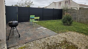 Une terrasse à l’arrière de la maison, avec un barbecue, deux chaises vertes, une clôture noire et un petit arbre.