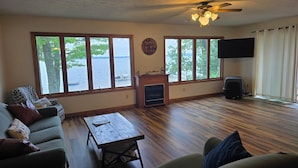 A living room with a sofa, a coffee table, a fireplace, a ceiling fan, and large windows overlooking a body of water.