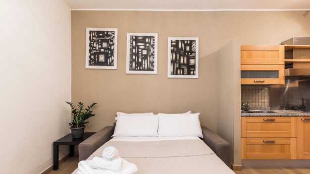 Une chambre d’hôtel comprenant un lit, une petite table avec une plante, un coin cuisine avec des placards en bois et trois œuvres d’art abstraites accrochées au mur.