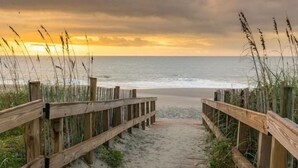Ein hölzerner Steg führt zu einem Strand, im Hintergrund ist ein Sonnenuntergang zu sehen.