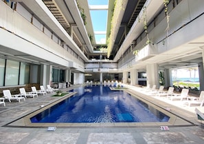 A modern indoor swimming pool with clear blue water, surrounded by white lounge chairs and a sun terrace with a view of the ocean.