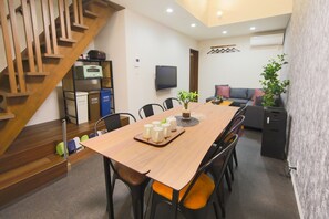 A modern living room with a wooden staircase, a dining area with a wooden table and chairs, a sofa, a TV, and a plant.