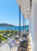 Un balcón con mesa y sillas, con vista a un puerto deportivo con barcos y un mar azul cristalino.