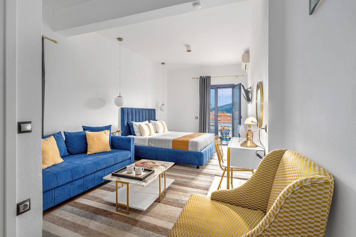 A modern hotel room with a blue sofa, a bed with a blue headboard, a yellow patterned armchair, and a view of the outdoors through a large window.