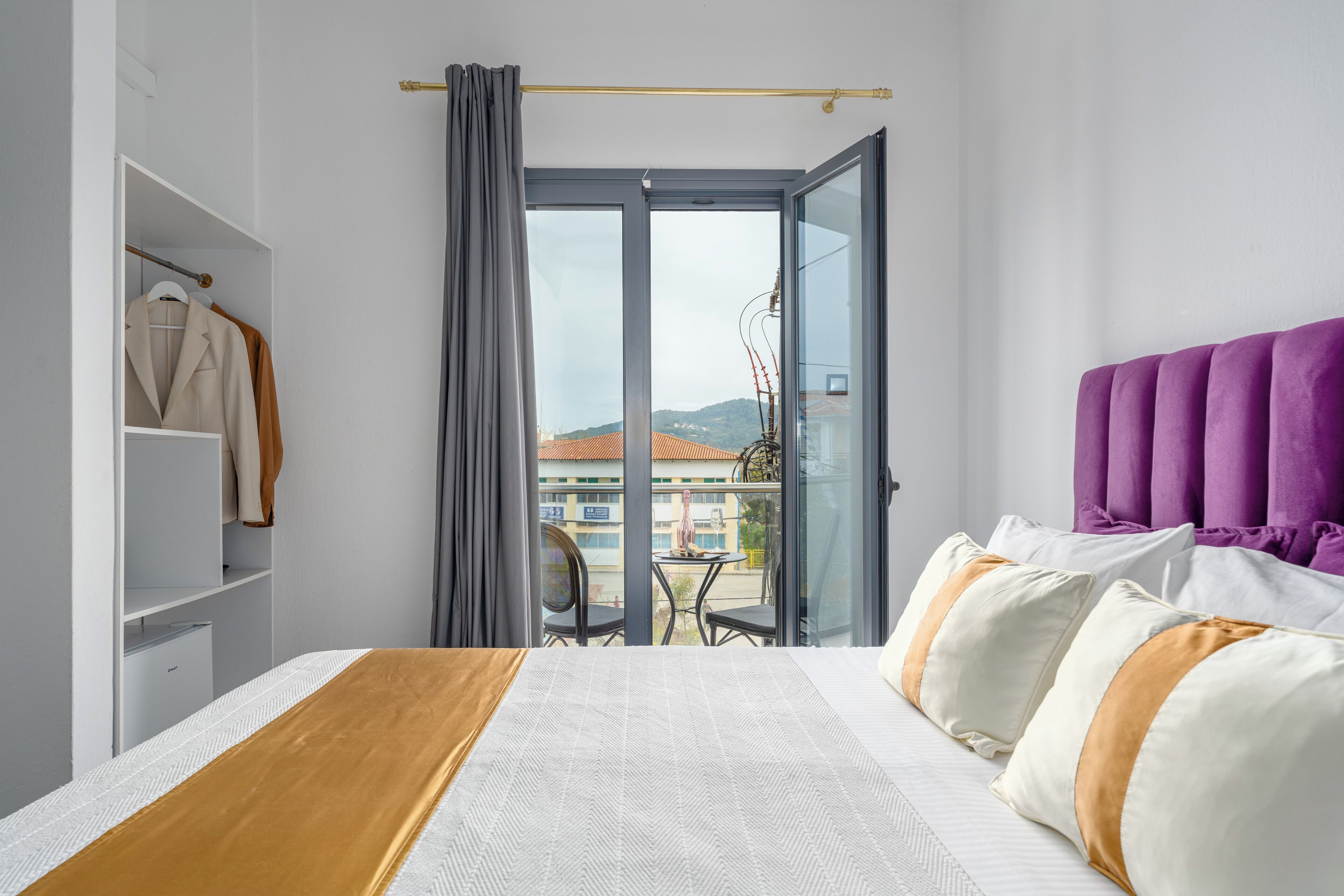 Habitación de hotel con cama, armario, balcón con mesa y sillas, y vistas a edificios y colinas.