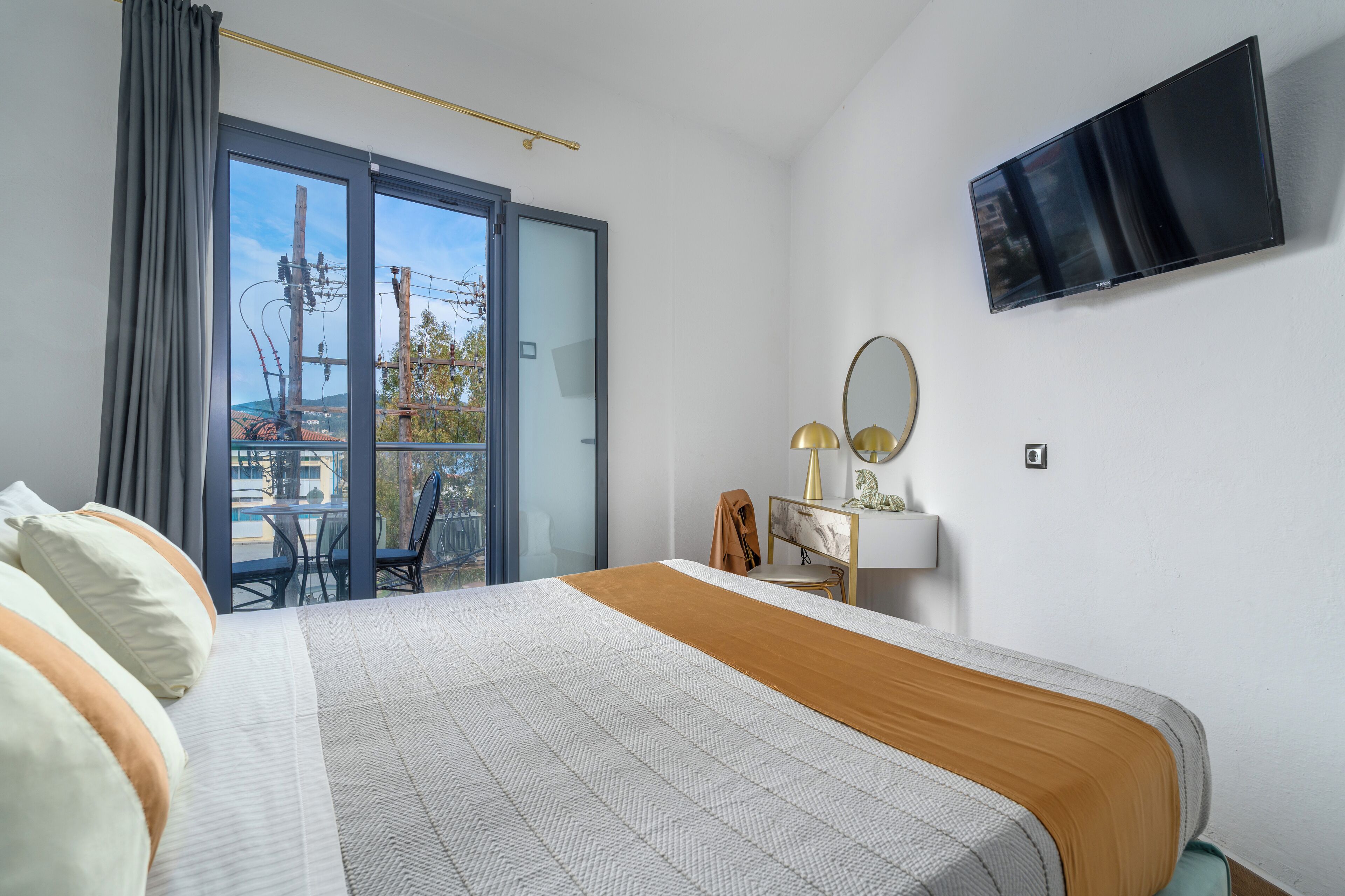 Habitación de hotel con cama, escritorio, silla, televisor y balcón con vistas a tendidos eléctricos y árboles.