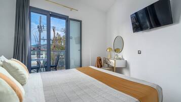 Habitación de hotel con cama, escritorio, silla, televisor y balcón con vistas a tendidos eléctricos y árboles.