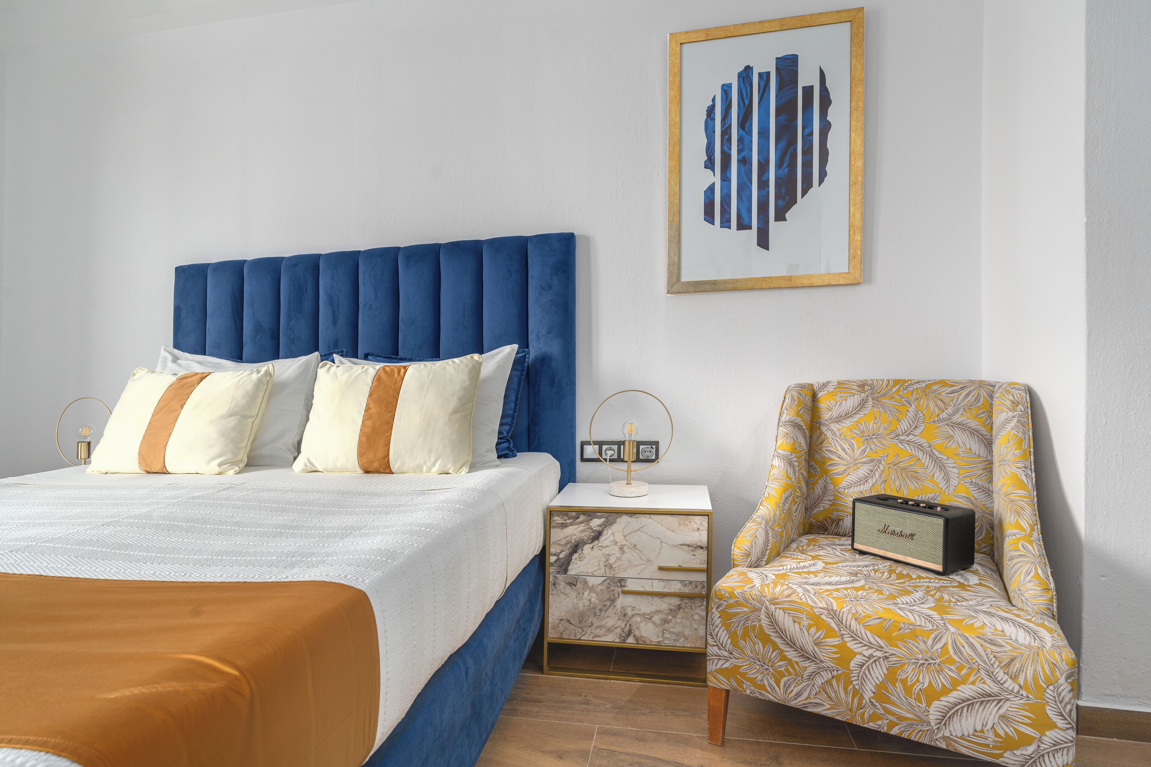 Un dormitorio con cabecera azul, una cama con ropa de cama blanca y naranja, una mesita de noche con un radio y una silla con estampado de hojas amarillas.