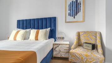 Un dormitorio con cabecera azul, una cama con ropa de cama blanca y naranja, una mesita de noche con un radio y una silla con estampado de hojas amarillas.