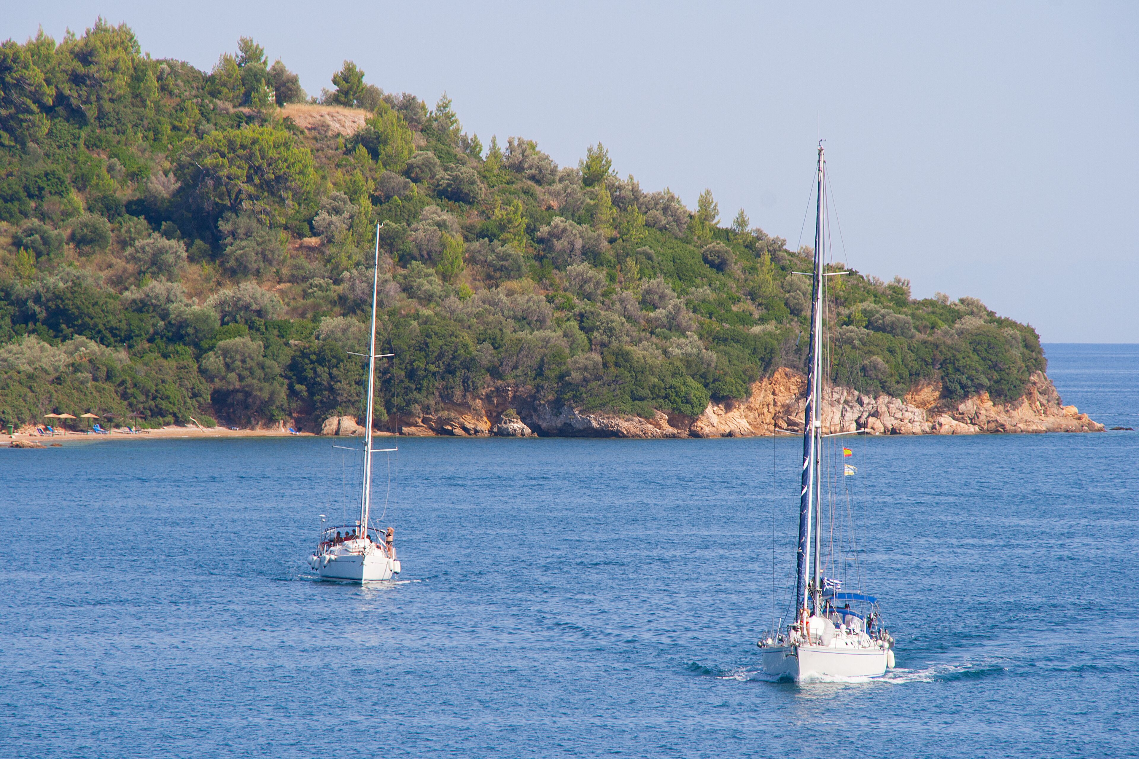 Two sailboats on a calm sea with a rocky coastline and dense greenery in the background.
