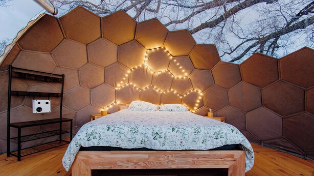 A bedroom with a hexagonal wall design, a wooden bed frame, and a bedside table with a lamp. - Les Bulles Perchées de Lartigue (Margouet-Meymes)