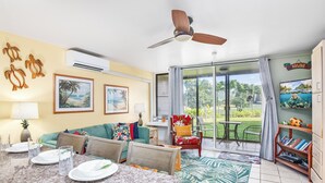 Un vaste espace de vie comprenant un coin repas avec une table, un ventilateur de plafond et une vue sur un paysage tropical grâce à de grandes fenêtres.