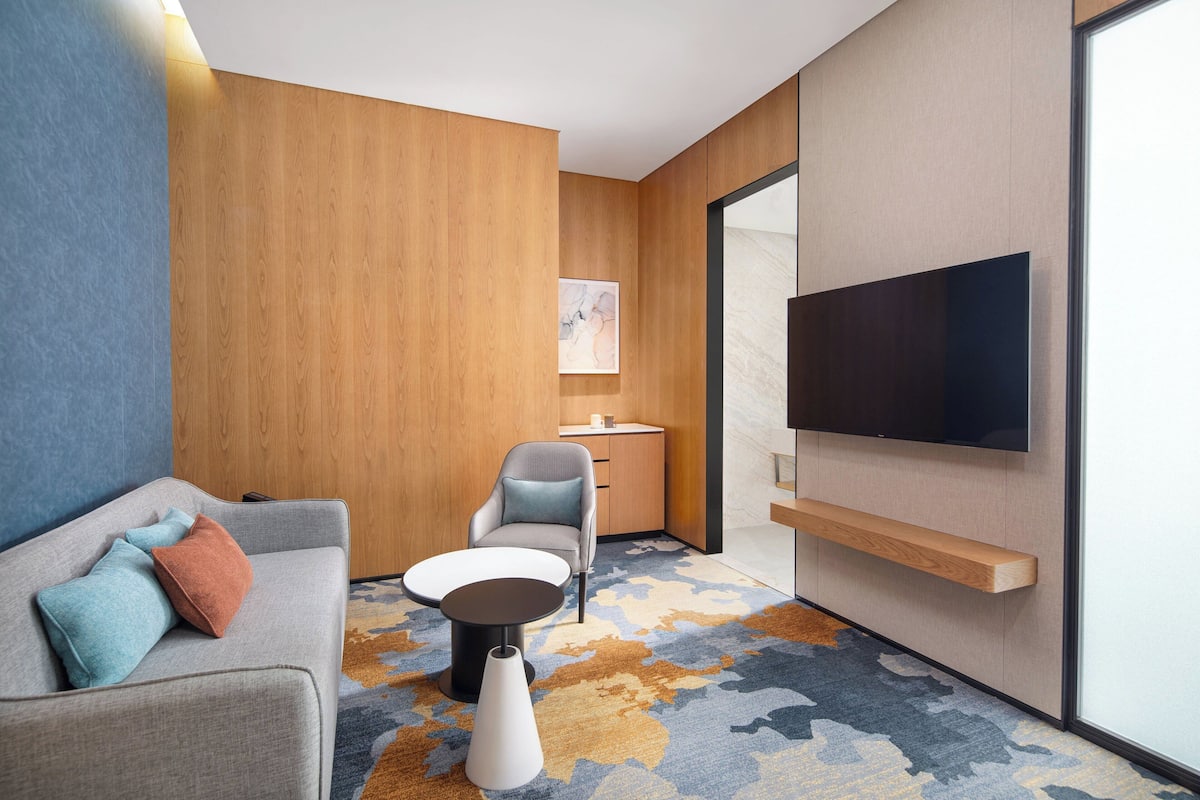 A modern hotel room with a grey sofa, a small round table, a grey armchair, a wooden cabinet, a flat-screen TV mounted on the wall, and a patterned carpet.